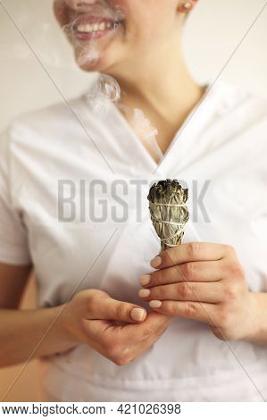 Unrecognizable Female Burning Sage Smudge Stick And Smelling Smoke Against Gray Wall During Meditati
