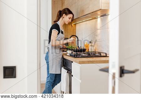 Young Woman In Apron Cooking Healthy Food At Modern Home Kitchen. Preparing Meal With Frying Pan On 