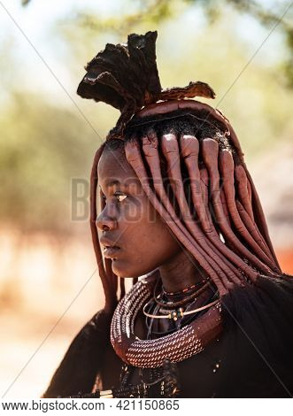 OPUWO, OTJIWARONGO REGIONNAMIBIA - MAY 4, 2021: Young unidentified Himba woman with the typical necklace and hairstyle in Himba living museum during a appearance in front of tourists