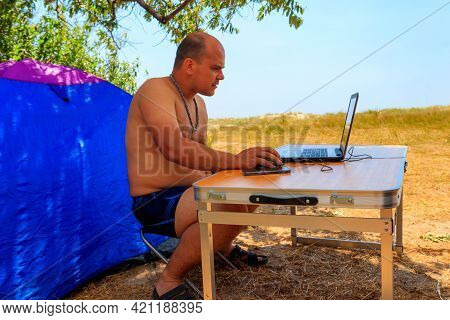 Man Using His Laptop Sitting At A Folding Picnic Table In A Camping. Working While Traveling. Freela