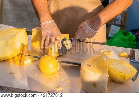 Shopkeeper's Hands Cutting A Head Of Cheese Close Up. Artisan Cheese In A Street Market. Cheese Make
