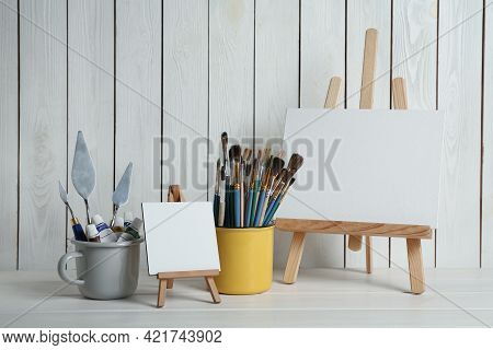 Easels With Blank Canvases, Paints And Brushes On White Wooden Table