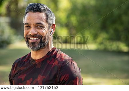 Mature fitness man listening to music while resting after jogging. Happy middle eastern man feeling refreshed after exercise. Portrait of a mixed race guy looking at camera in park after workout.