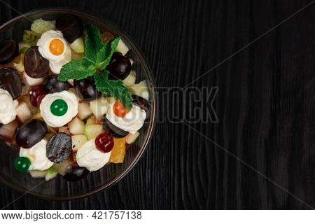 Salad from grapes apples pears kiwi oranges with mascarpone chease and cream. Healthy fresh fruit summer salad in glass bowl on black wooden background with copy space.