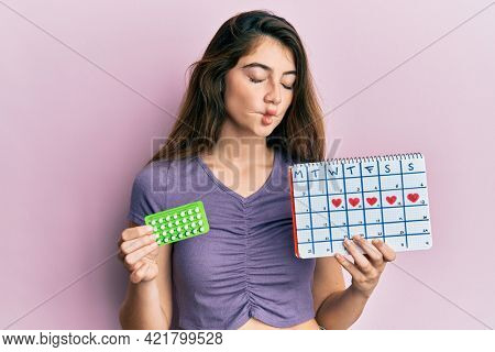 Young caucasian woman holding birth control pills and menstrual calendar making fish face with mouth and squinting eyes, crazy and comical. 