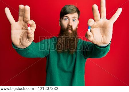 Redhead man with long beard holding two different pills making fish face with mouth and squinting eyes, crazy and comical. 