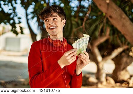 Young hispanic man using headphones counting argentinian pesos at the city