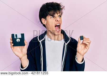 Handsome hipster young man holding floppy disk and sdxc card angry and mad screaming frustrated and furious, shouting with anger. rage and aggressive concept. 