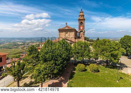 View of old catholic church among green trees under beautiful blue sky with white clouds in small town of Diano d'Alba, Piedmont, Northern Italy.