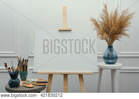 Easel With Blank Canvas, Brushes, Paints And Palette In Studio