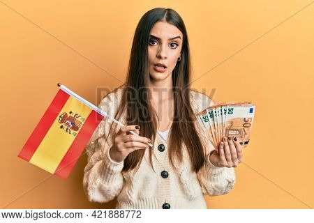 Beautiful brunette young woman holding spain flag and euros banknotes in shock face, looking skeptical and sarcastic, surprised with open mouth 
