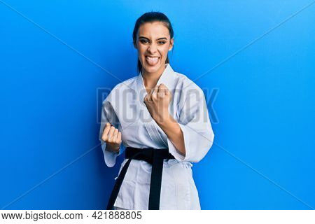 Beautiful brunette young woman wearing karate fighter uniform with black belt doing attack pose sticking tongue out happy with funny expression. 