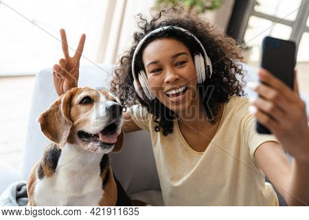 Happy young african woman in headphones relaxing on a couch with her pet dog at home, taing a selfie