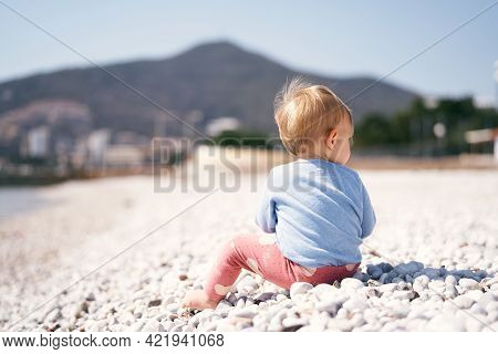 Kid In A Blue Sweater And Red Pants Sits With His Head Turned To The Right On A Pebble Beach Against