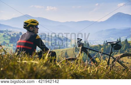 Young Man In Safety Helmet And Glasses Sitting On Grass Near Bicycle. Male Bicyclist Resting On Gras