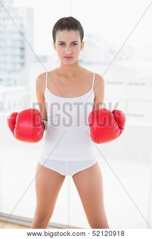 Unsmiling natural brown haired woman in white sportswear wearing boxing gloves in bright living room