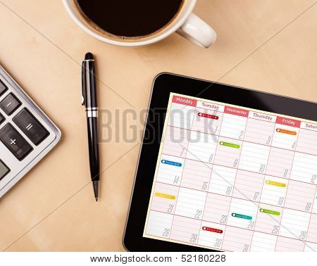 Workplace with tablet pc showing calendar and a cup of coffee on a wooden work table close-up