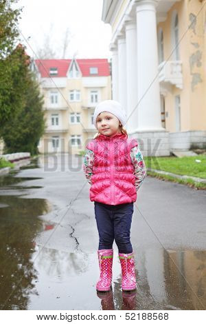 Cute girl in bright clothes standing in a puddle on a cloudy day