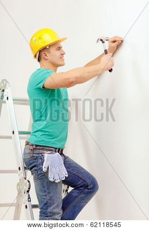 reapir, building and home renovation concept - smiling man in yellow protective helmet hammering nail in wall