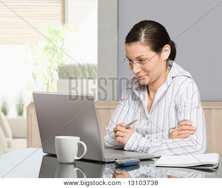 Jonge vrouw zit aan bureau werken met laptopcomputer thuis, glimlachend.