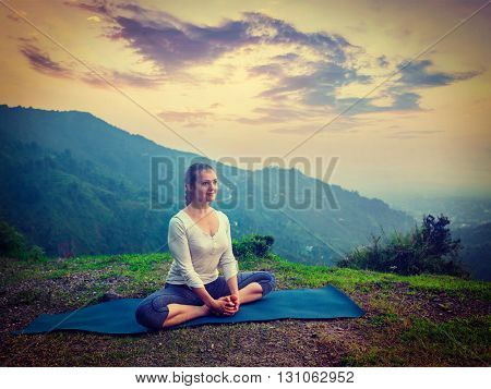 Vintage retro effect hipster style image of sporty fit woman practices yoga asana Baddha Konasana - bound angle pose outdoors in HImalayas mountains on sunset. Himachal Pradesh, India
