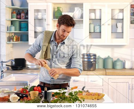 Young man preparing food at home in kitchen using pepper-mill.