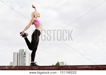 Young blonde woman is doing leg stretching outdoors warming up before training pulling the hip on a background of clouds