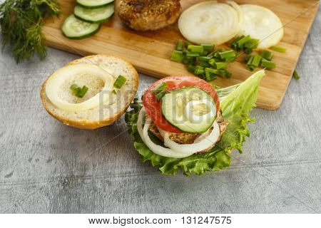 Cooking process of a sandwich burger ingredients on wooden cutting board on wooden table against white background fresh vegetables herbs fried meat buns sauces and knife horizontal view shallow DOF
