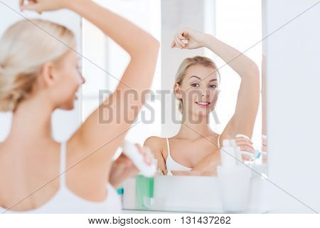 beauty, hygiene, morning and people concept - smiling young woman applying antiperspirant or stick deodorant and looking to mirror at home bathroom