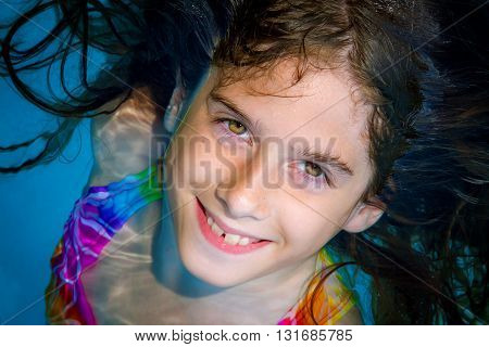 A tween girl looks up from a swimming pool and smiles with big eyes and buck teeth.