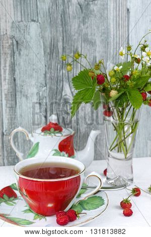 Fresh wild strawberry tea in an colorful cup and teapot and bouquet of ripe wild strawberries in a vase on wooden background