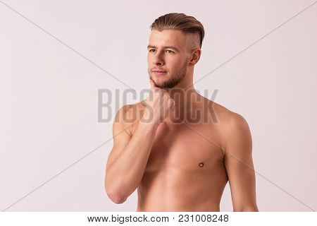 Studio shot of handsome bearded hipster touching chin and looking away while standing isolated on white background. Young masculine man posing against white background. Men's beauty and skin care