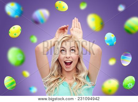 easter, holidays and people concept - happy smiling young woman making bunny ears over ultra violet background with colored eggs
