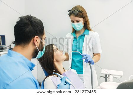 Smiling Beautiful Patient Sitting In Chair With Dental Purple Bib, Male Dentist, In Blue Uniform And