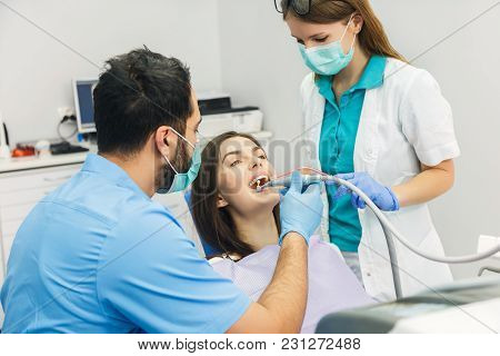 Smiling Beautiful Patient Sitting In Chair, Male Dentist, In Blue Uniform And Gloves, Holding Explor