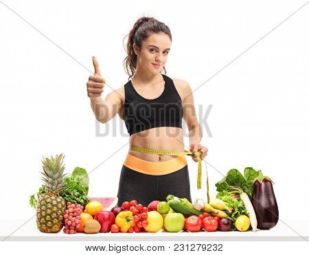Fitness girl measuring her waist with a measuring tape and making a thumb up sign behind a table with fruit and vegetables isolated on white background