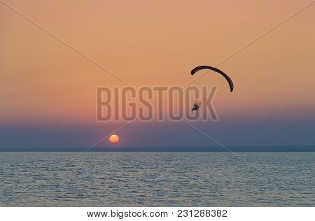 Silhouette Of Powered Paraglider Soaring Flight Over The Sea Against Marvellous Orange Sunset Sky. P