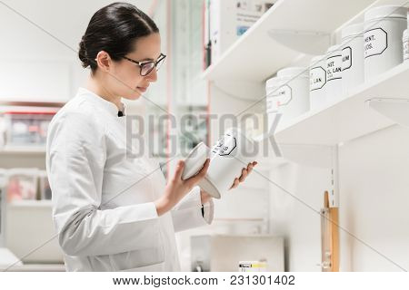 Side view of an experienced female pharmacist checking the container of a chemical pharmaceutical substance during inventory in a modern drugstore