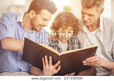 Generation Portrait. Grandfather, Father And Son Sitting And Reading A Book On Sofa.