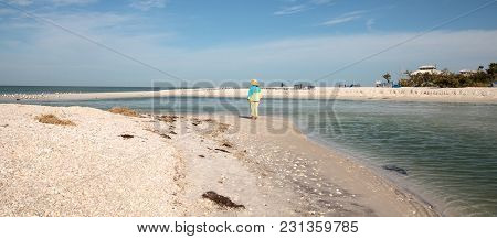 Older Woman Stands On The White Sand Beach In Front Of Aqua Blue Water Of Clam Pass