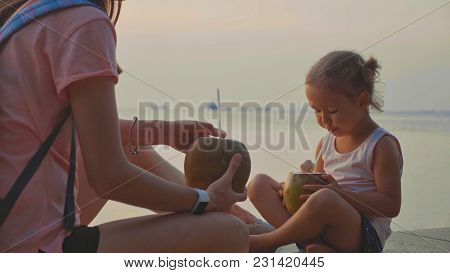 Travelers Mother And Little Daughter Sits At Seafront And Drinks Coconats In Trip. Portrait Of Cute 