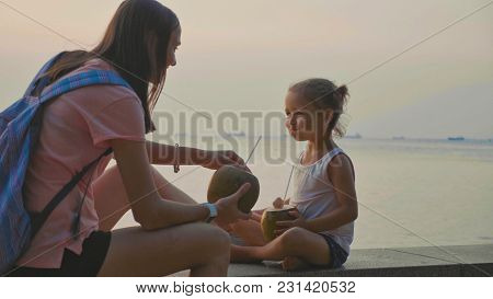 Travelers Mother And Little Daughter Sits At Seafront And Drinks Coconats In Trip. Mom Shares Her Co