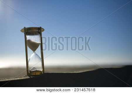 Backlit Sand Timer On The Peak Of A Sand Dune, With Sand And Sky Behind It.