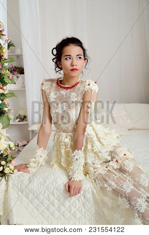 Asian-american Girl Model In Beautiful Dress Sitting On A White Bed With Flowers And Looks Up. Portr
