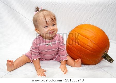 A Baby Girl With Her Hair Tied In A Ribbon On Top Of Her Head Sits Straddling On A White Blanket Nex