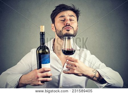 Concentrated Man Holding Bottle Of Wine And Smelling Beverage With Eyes Closed On Gray Backdrop.