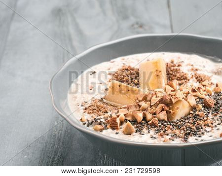 Close Up View Of Overnight Oats In Bowl With Banana, Lsa, Chia Seeds, Almond And Honey On Gray Woode