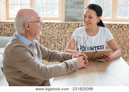 Social Volunteering. Merry Optimistic Volunteer Carrying Book While Talking To Senior Man And Lookin