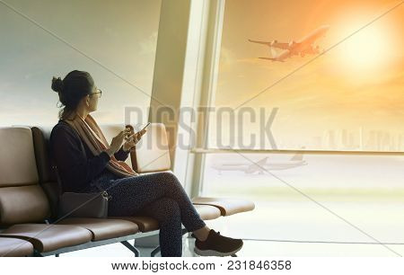 Traveling Woman Sitting In Airport Terminal With Smart Phone In Hand Looking Through Window To Flyin