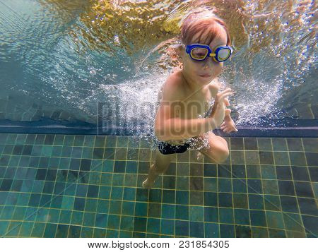 A Child Boy Is Swimming Underwater In A Pool, Smiling And Holding Breath, With Swimming Glasses.